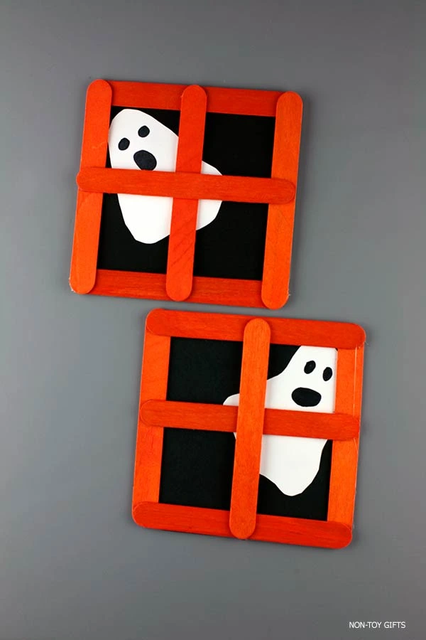 Halloween kids craft featuring a ghost peeking through an orange popsicle stick window frame on black paper background.