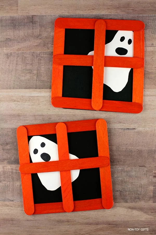 Easy Halloween craft for kids showing a white ghost cutout peeking out from behind an orange popsicle stick window glued on black paper
