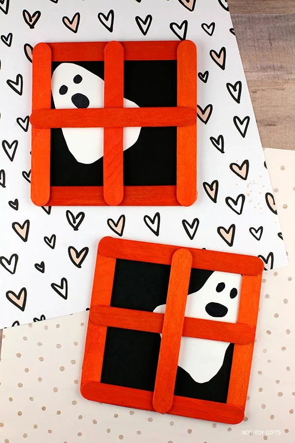 DIY Halloween ghost window craft made with orange-painted craft sticks, black paper background, and a ghost cut from white paper with drawn face.